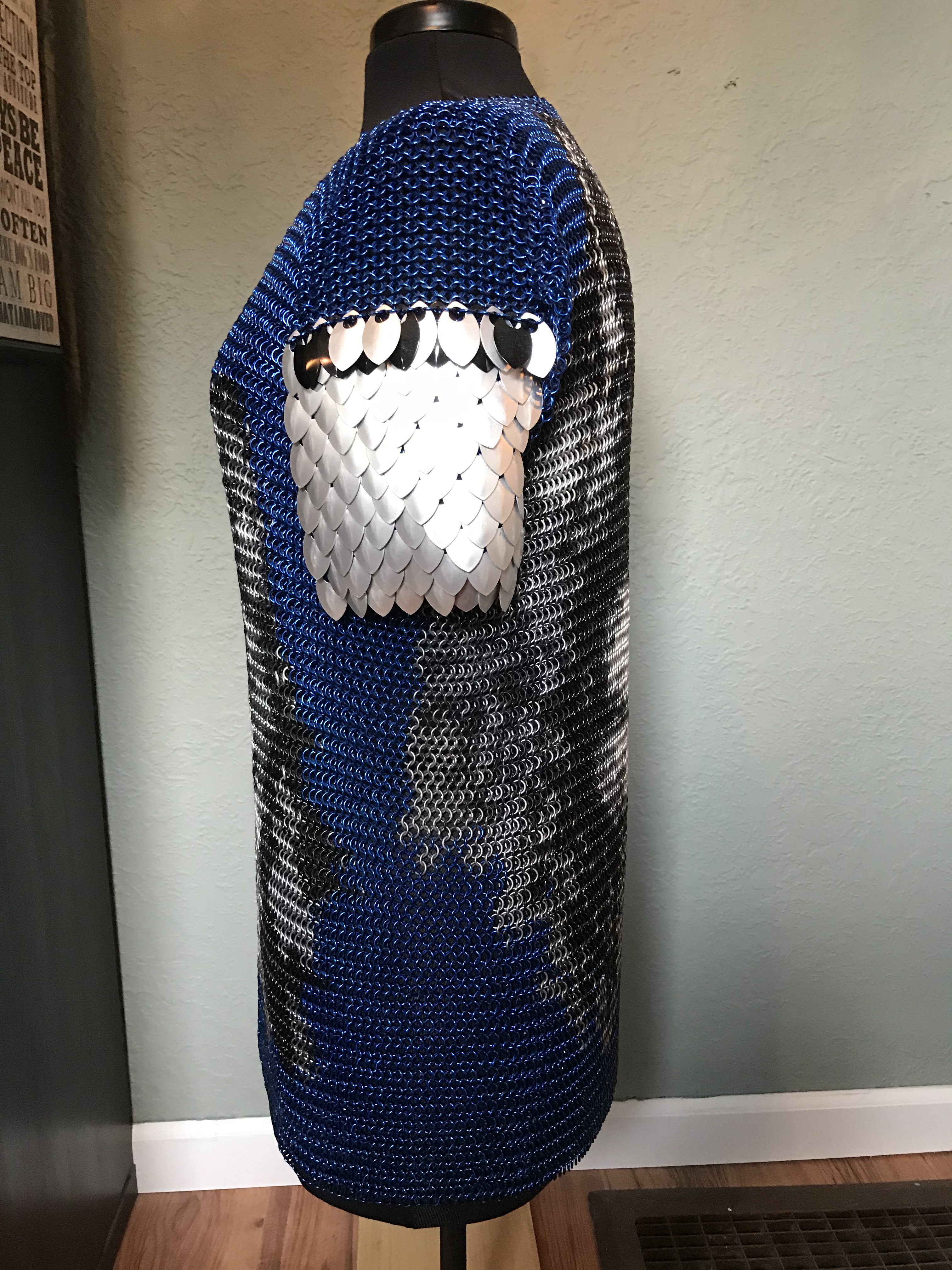 a chainmaille shirt with a detailed wolf’s face in black, silver, and gold rings on a deep blue background, with silver scale embellishments on the shoulders.
