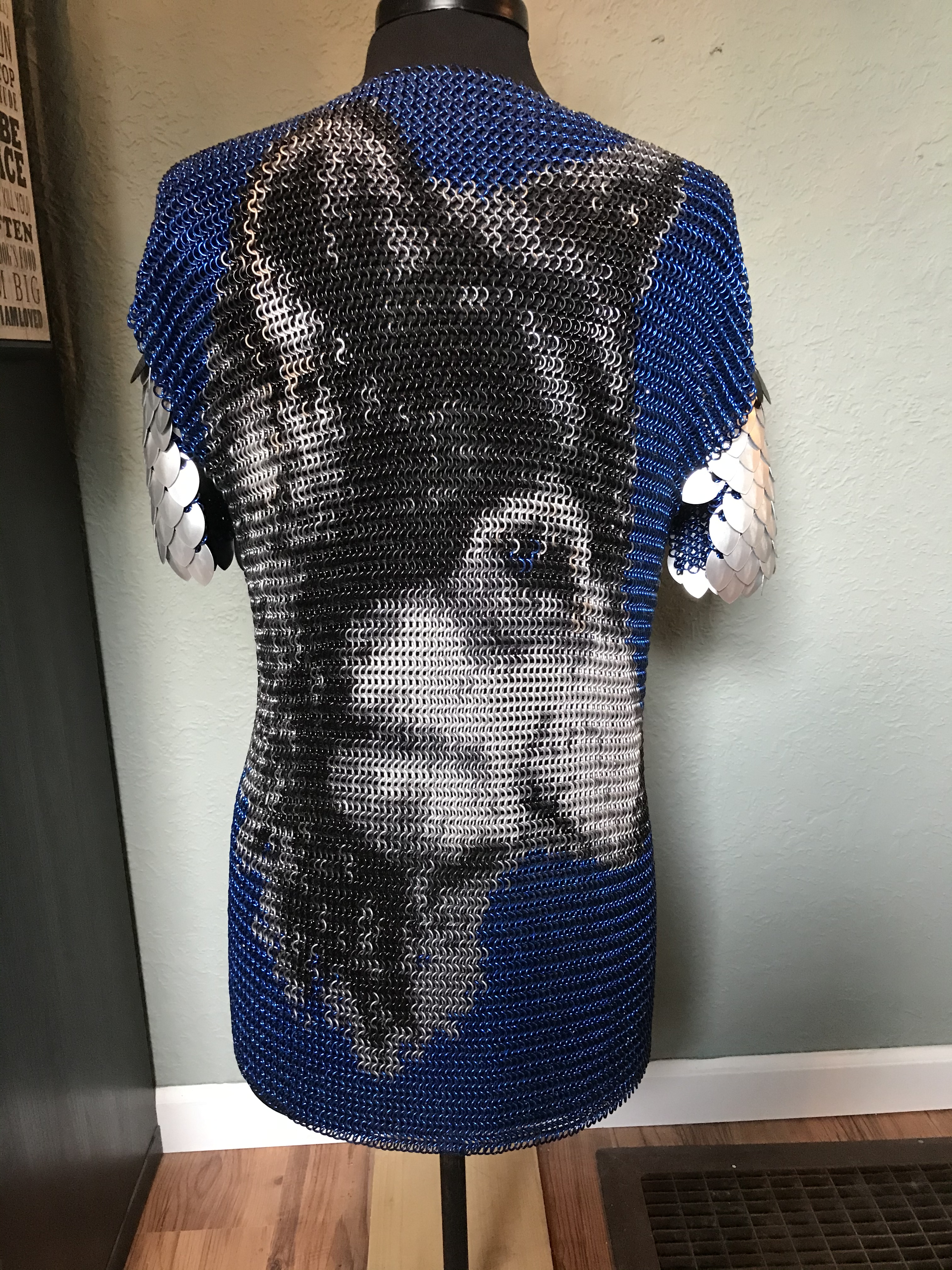 a chainmaille shirt with a detailed wolf’s face in black, silver, and gold rings on a deep blue background, with silver scale embellishments on the shoulders.

