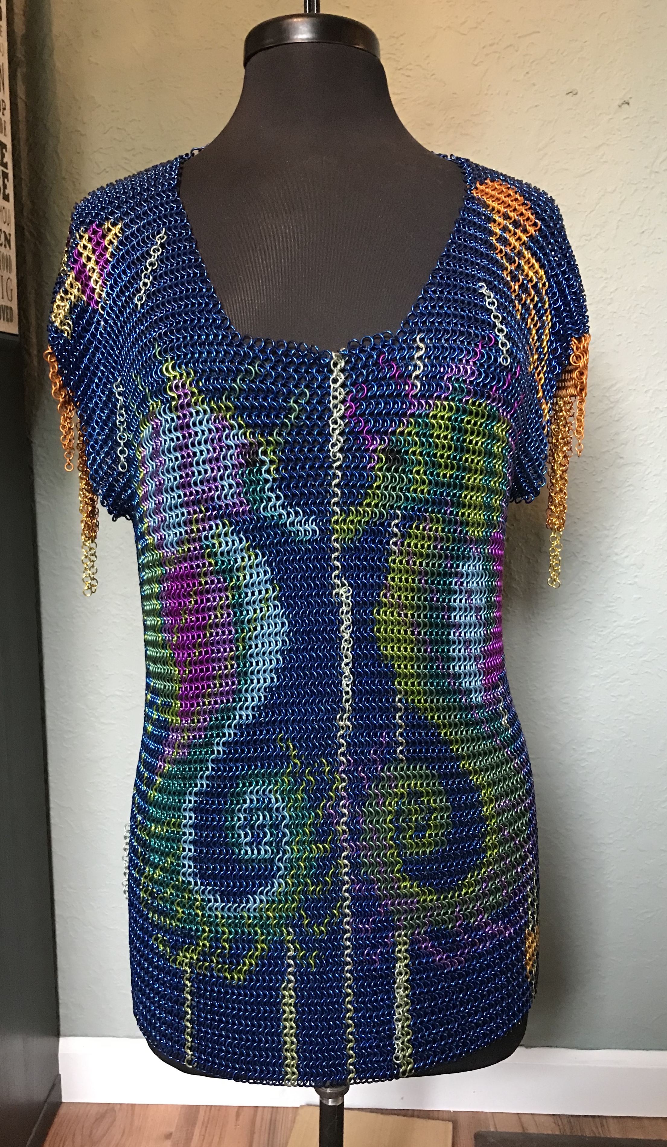 a chainmaille shirt with two colorful seahorses facing each other in pink, purple, blue, green, and yellow rings on a deep blue background, with bronze chain fringe sleeves.
