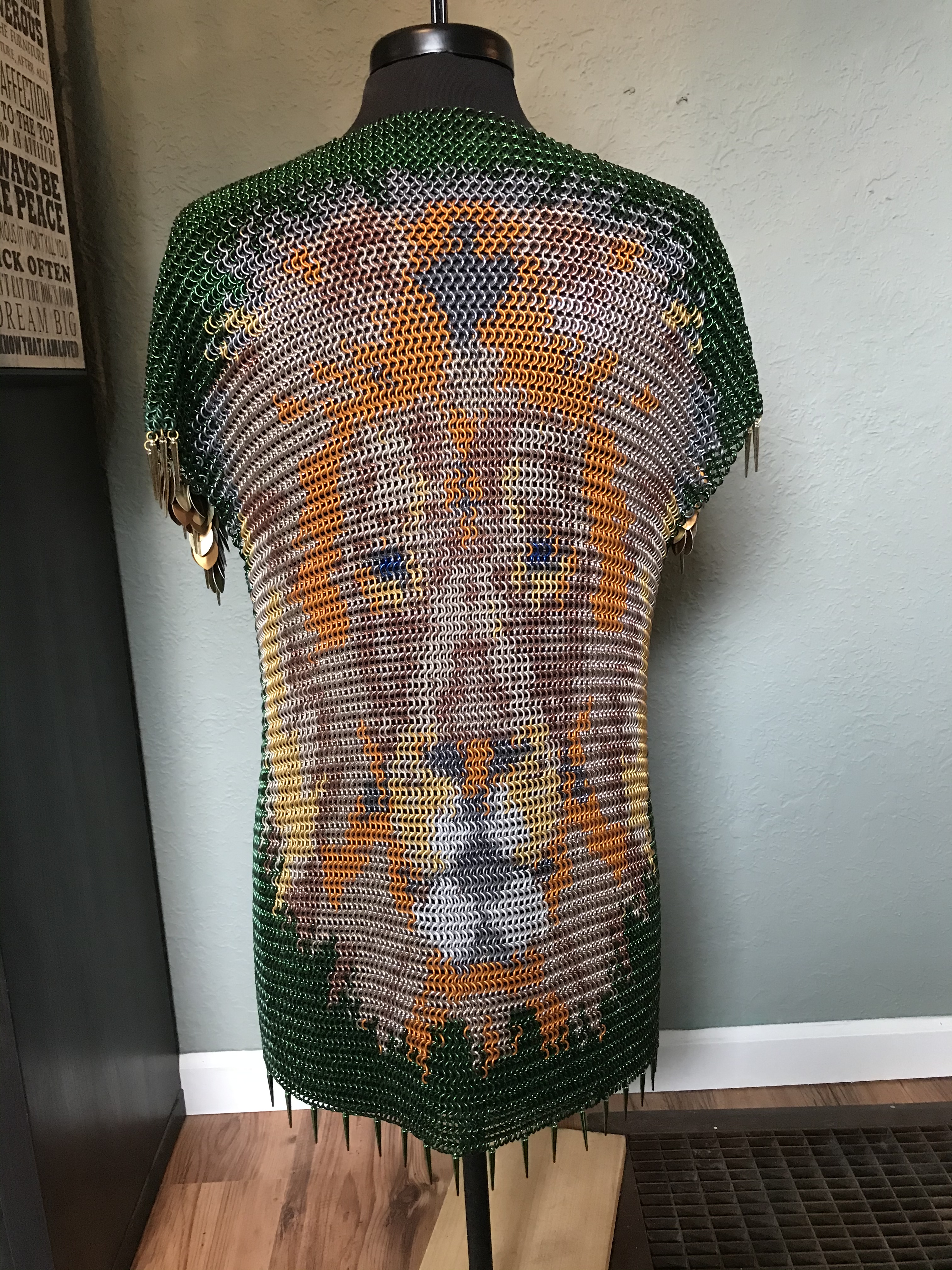 A chainmaille shirt, featuring a detailed lion’s face in colored rings with a gold crown, on a forest green background.