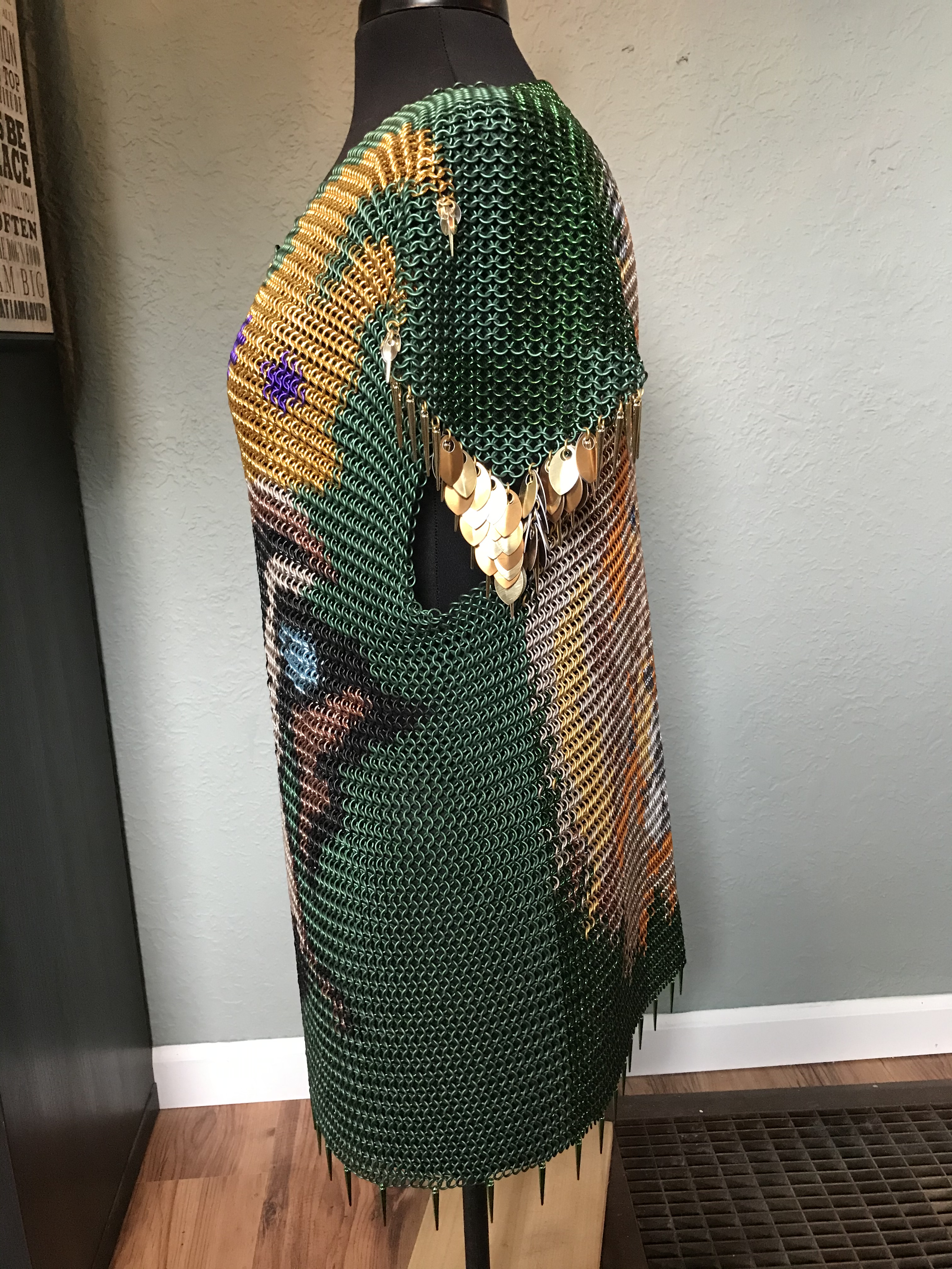 A chainmaille shirt, featuring a detailed lion’s face in colored rings with a gold crown, on a forest green background.