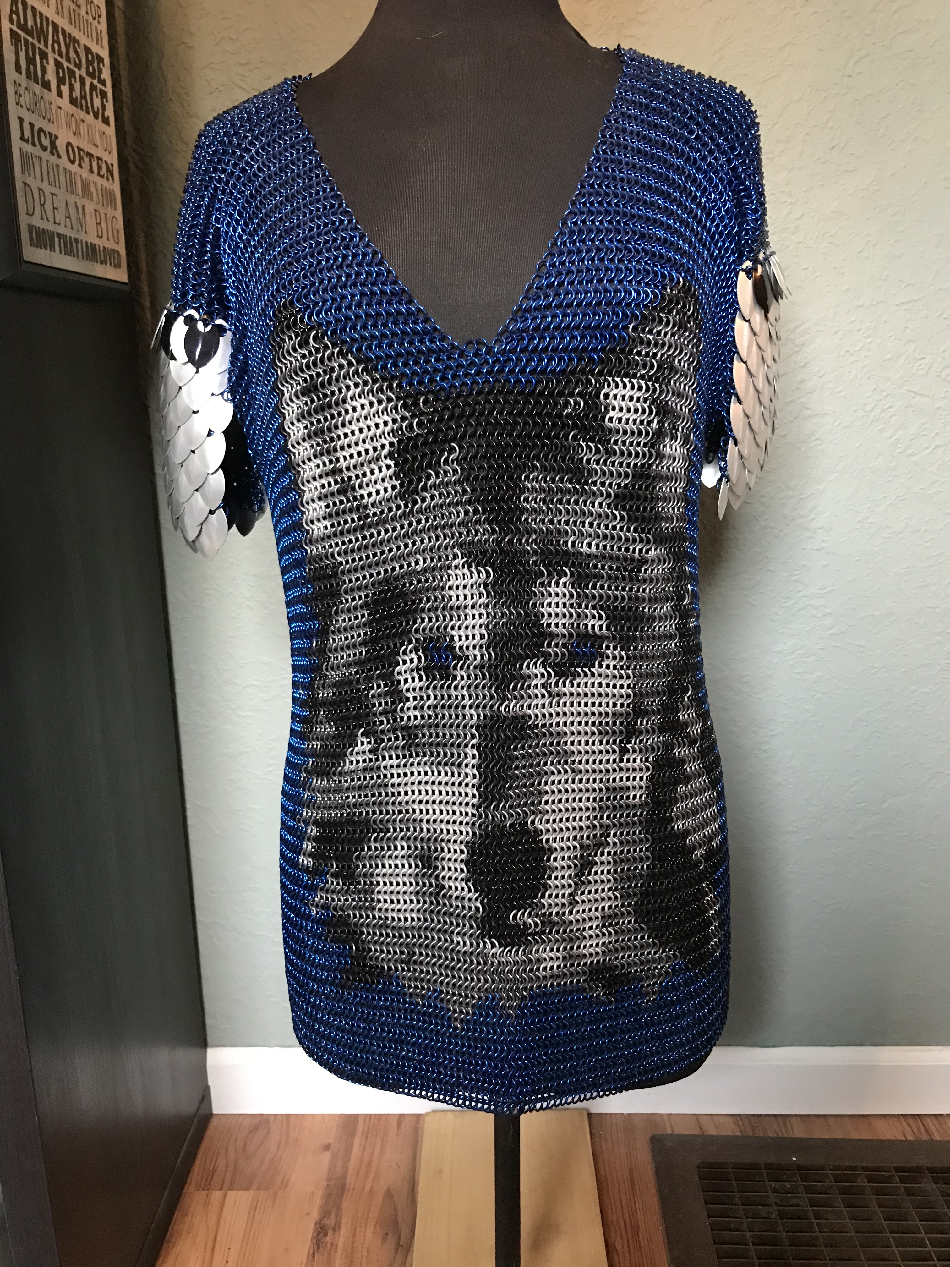 a chainmaille shirt with a detailed wolf’s face in black, silver, and gold rings on a deep blue background, with silver scale embellishments on the shoulders.

